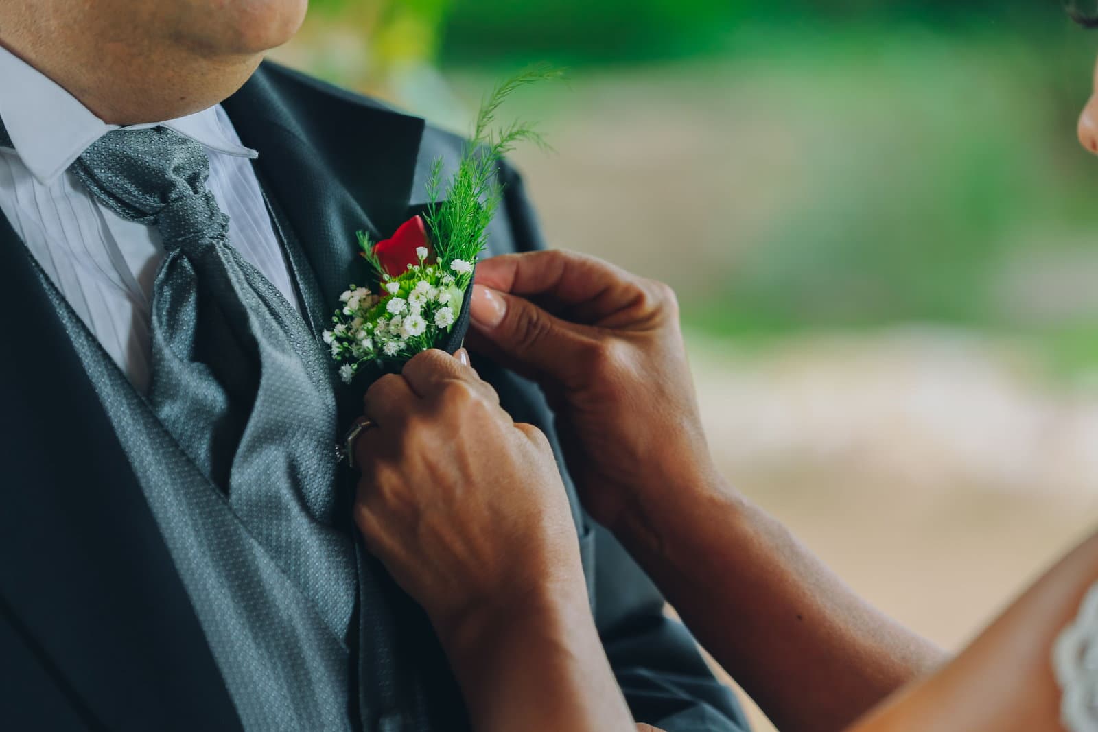 Hands pin a white and red boutonniere to the lapel of a man in a grey morning suit and cravat outdoors.