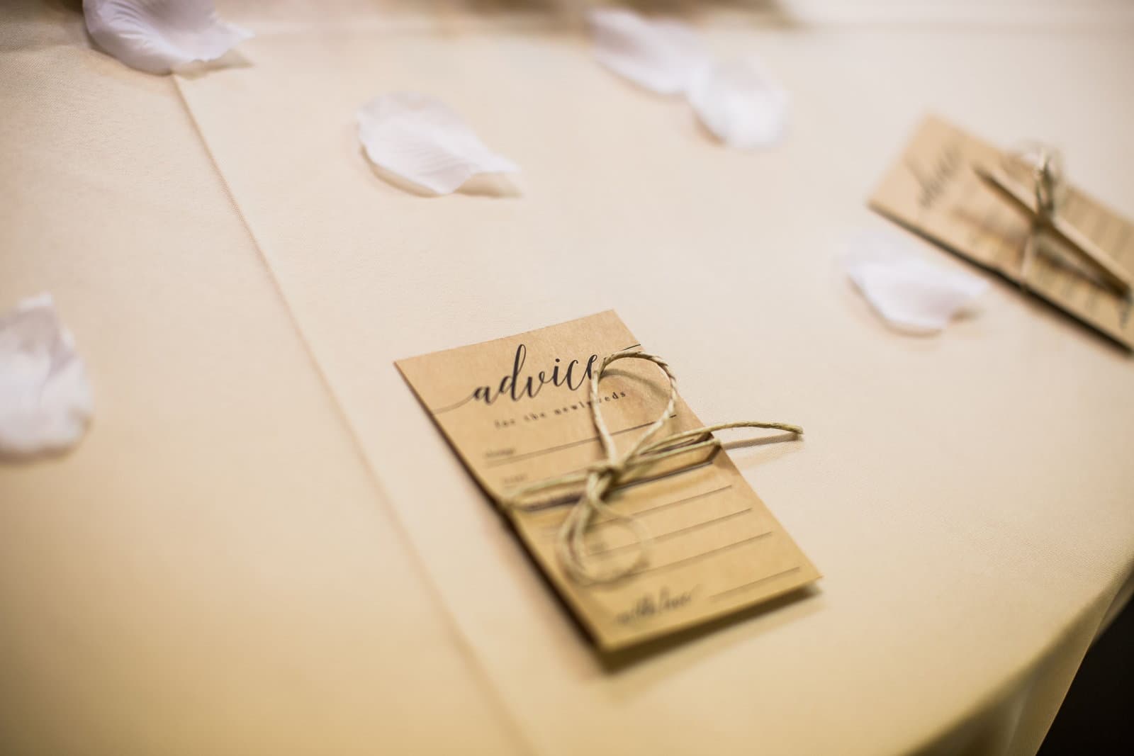 An advice card for the newlyweds, tied with twine, rests on a cream wedding table scattered with white petals.