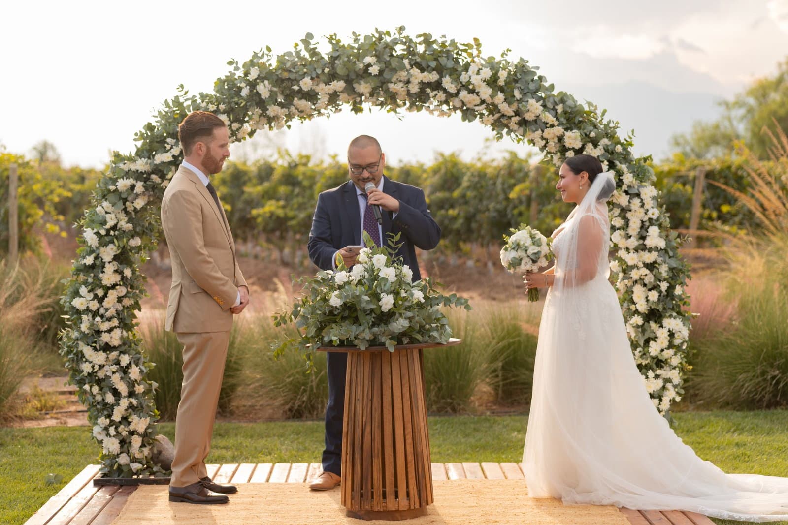 A friend in a navy suit officiates from a wooden lectern between the couple under a floral arch outdoors.