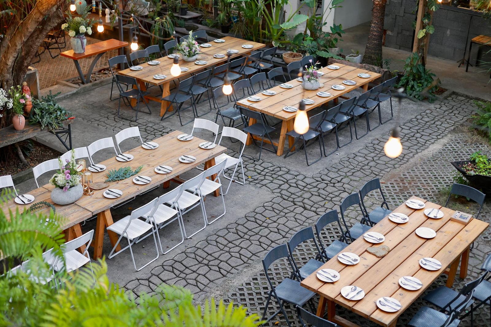 An overhead view of long wooden rehearsal dinner tables set for guests in a courtyard strung with bulb lights.