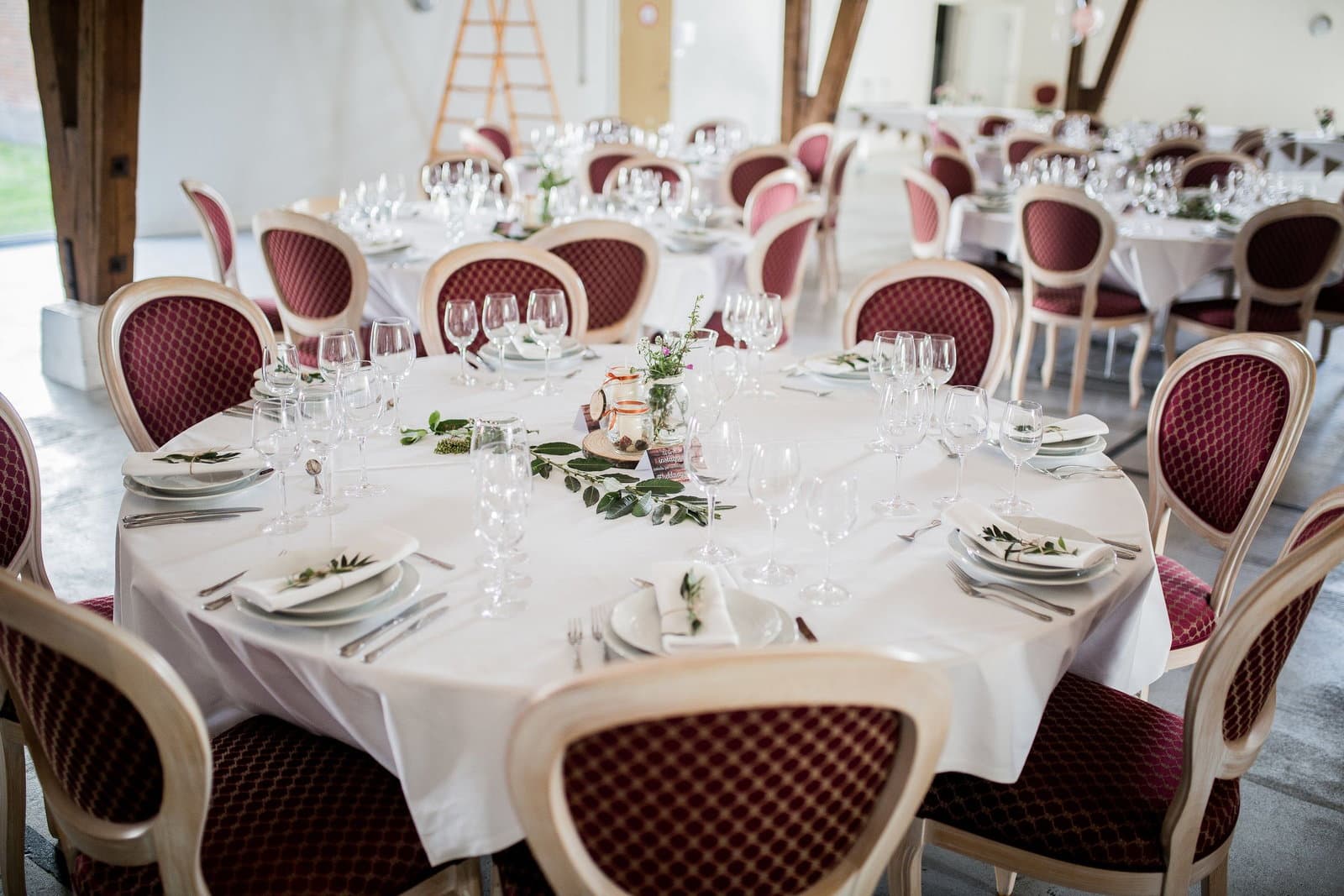 Round reception tables laid with glassware and greenery, surrounded by upholstered chairs in a bright venue.