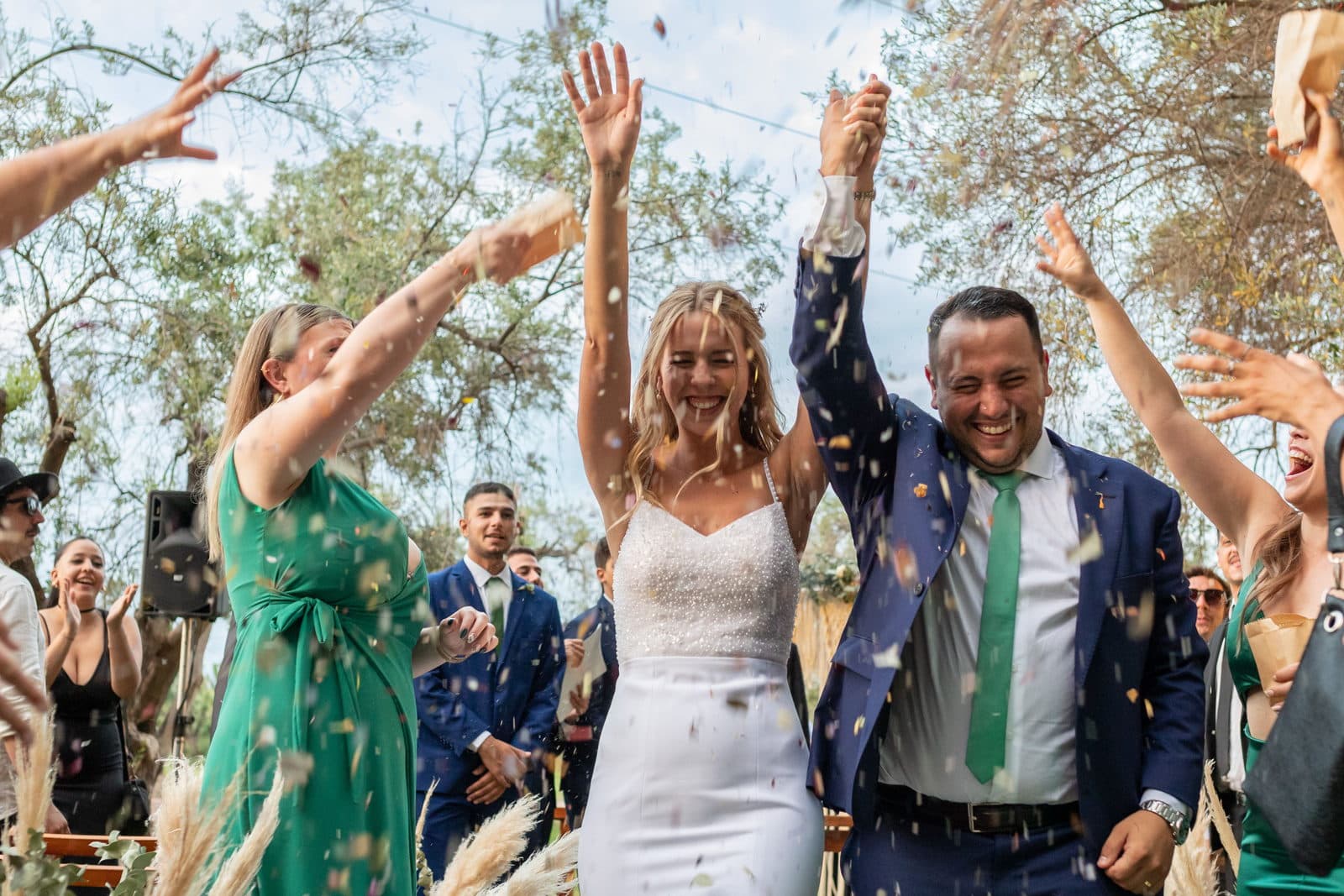 Guests in green dresses and navy suits throw confetti over the couple as they walk out of a daytime garden ceremony.