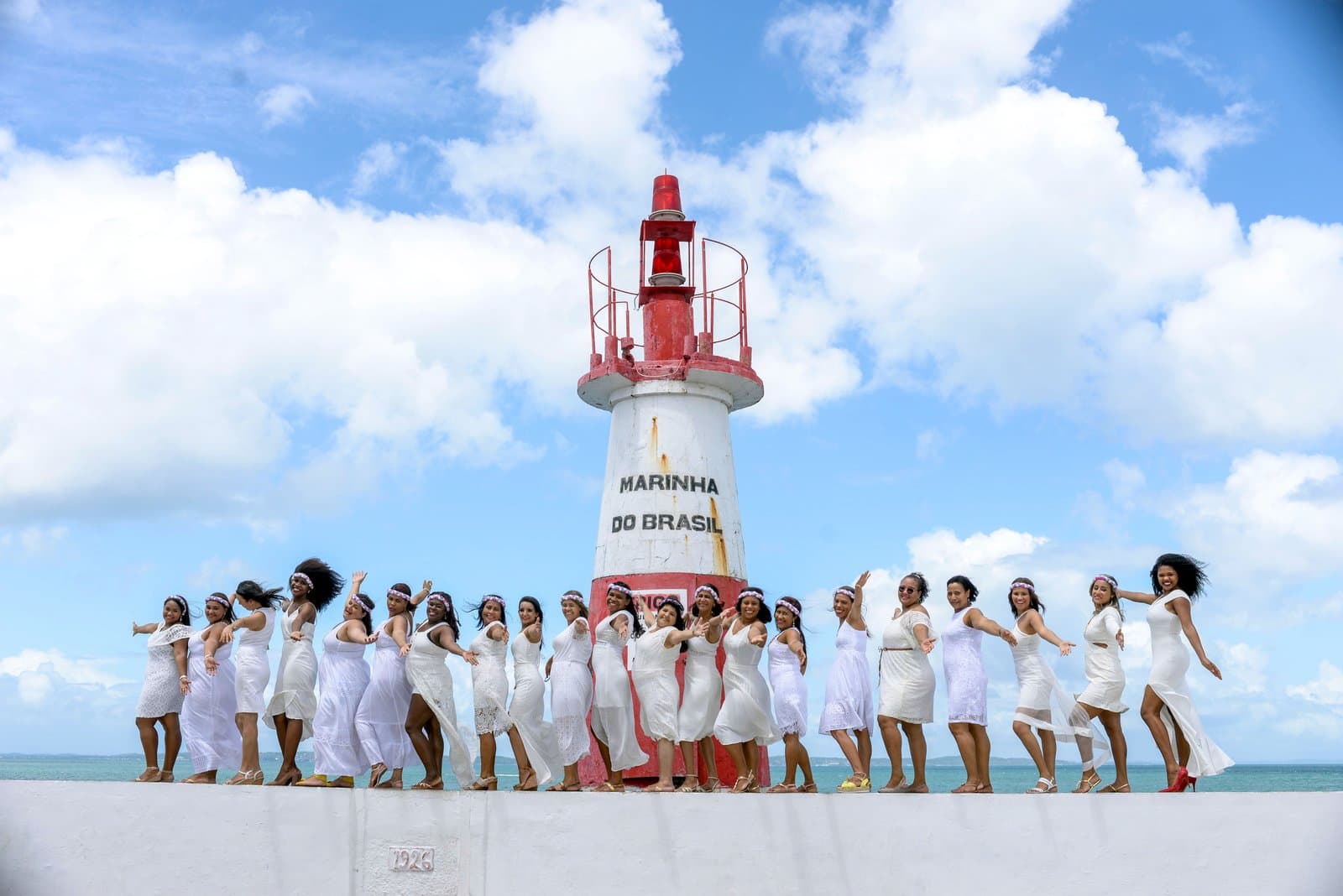 A large bachelorette group in white dresses poses along a seawall beside a red and white lighthouse under a bright blue sky.