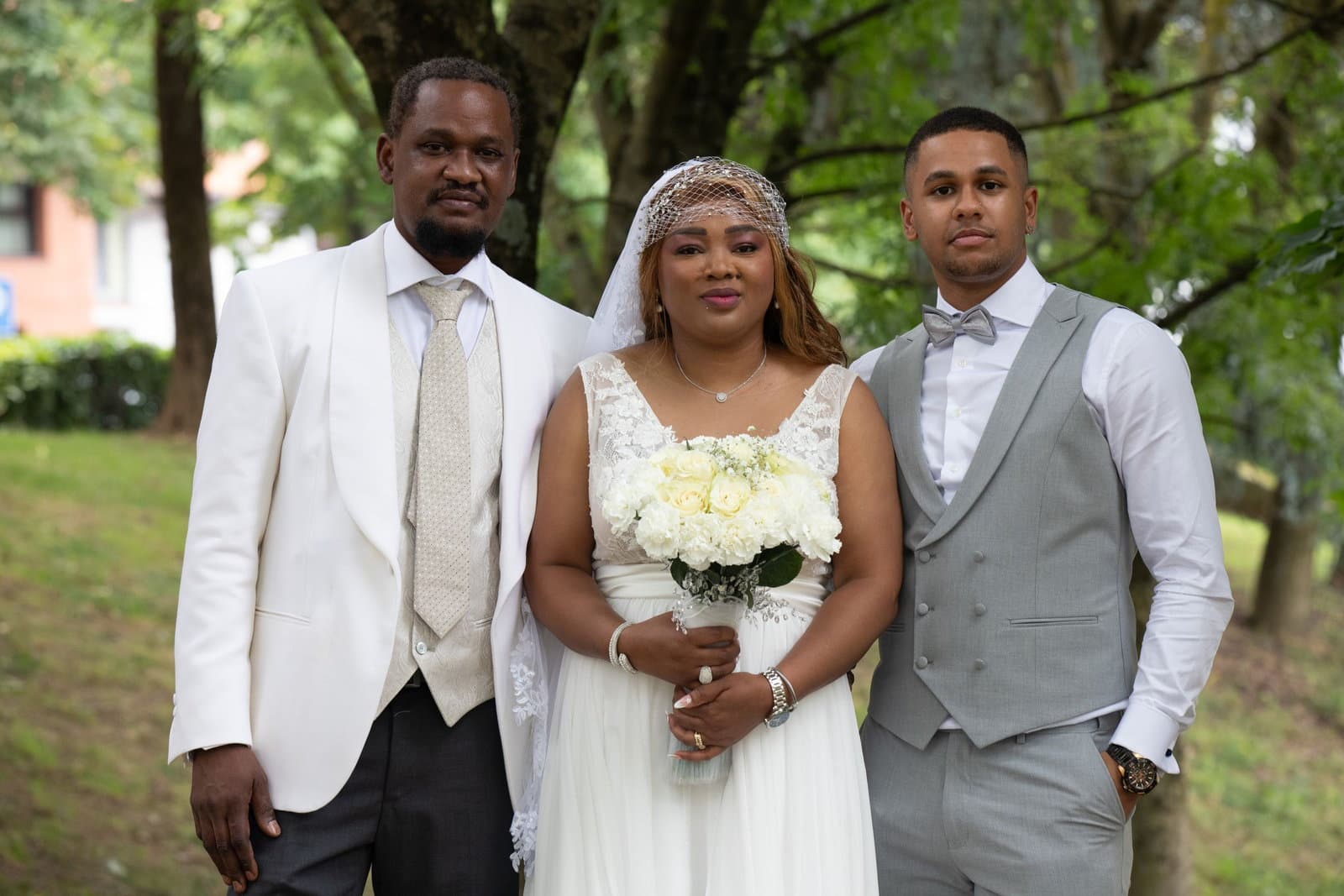 A bride stands between the groom and his best man in matching suits on a sunlit lawn after the ceremony.