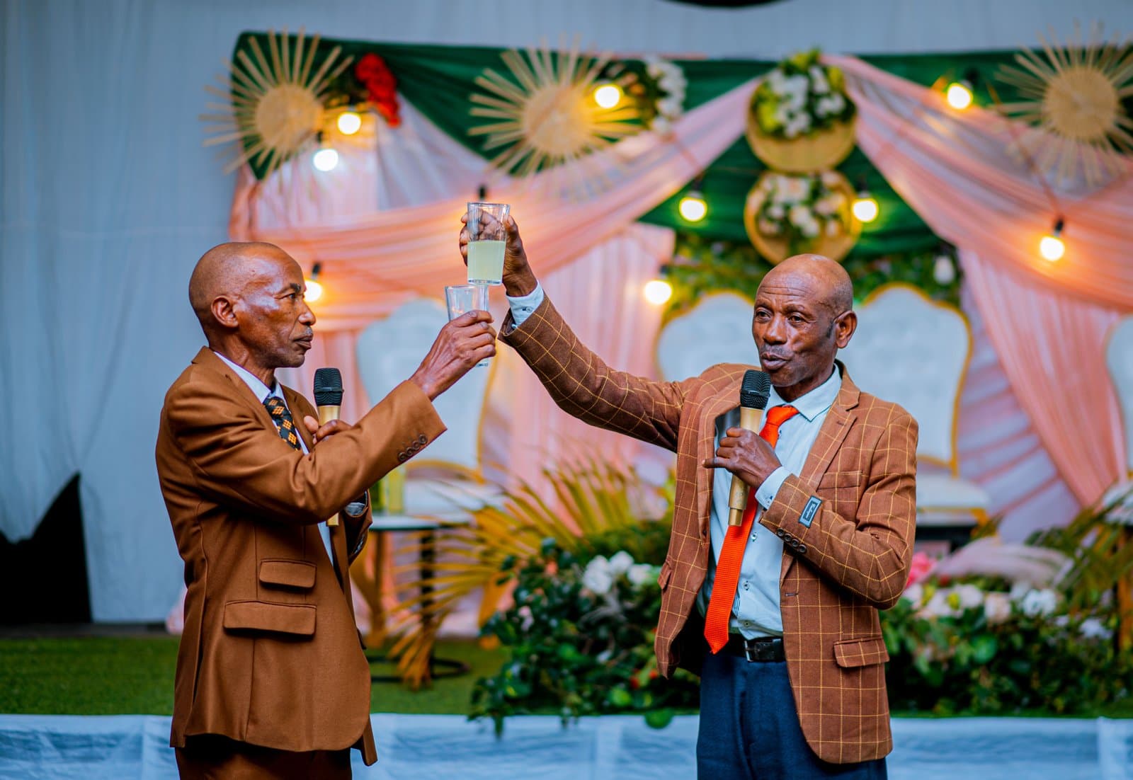 Two men in suits hold microphones and raise their glasses to each other during a toast at a decorated wedding marquee.