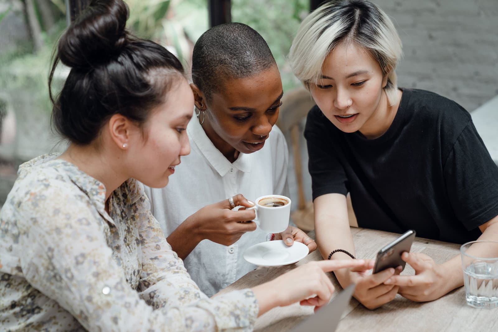 Three friends lean over one phone at a cafe table, one pointing at the screen beside a cup of coffee.
