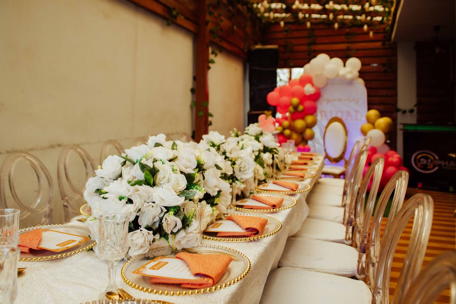 A long bridal shower table set with white roses, gold-rimmed plates and a pastel balloon arch.