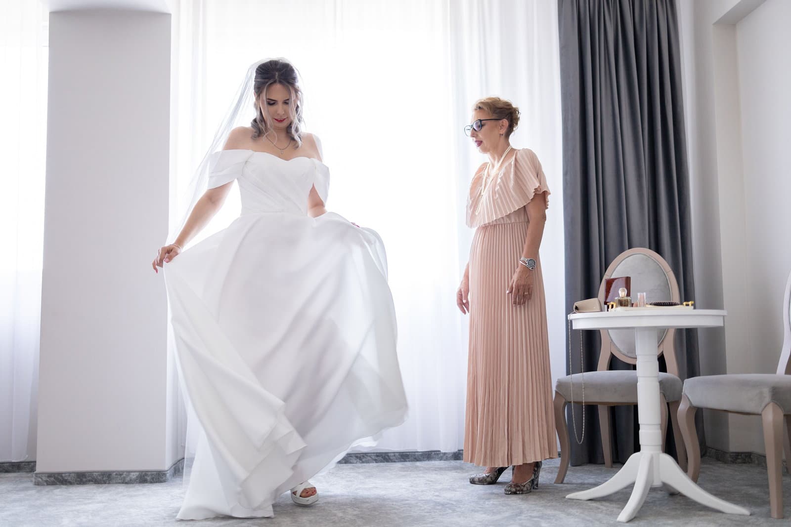 A bride sweeps her gown out in a bright white bridal suite while her attendant in a blush dress watches from the window.