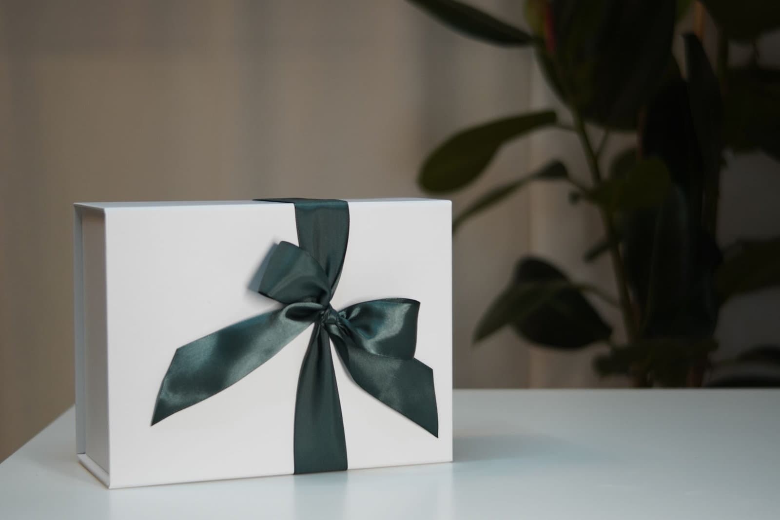 A white gift box tied with a wide dark green satin ribbon sits on a pale table beside a houseplant.