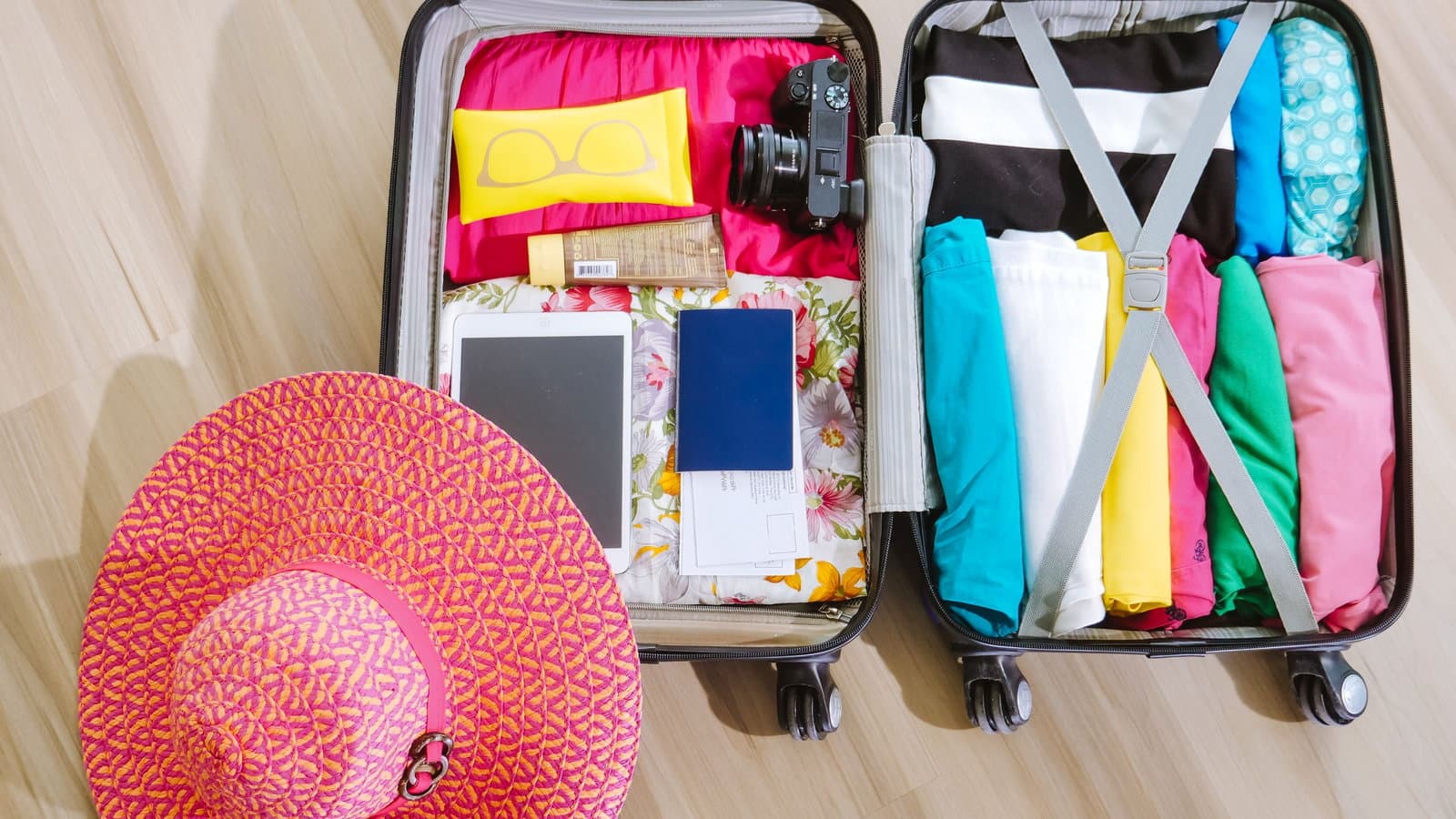 An open suitcase packed with neatly folded clothes, a passport, sunglasses and a sun hat, photographed from above.