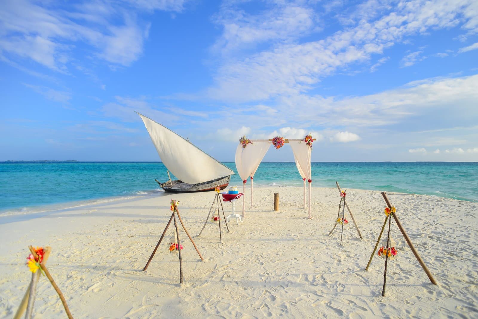 A flower-trimmed wedding arch on white sand beside a traditional sailing boat and bright turquoise water.