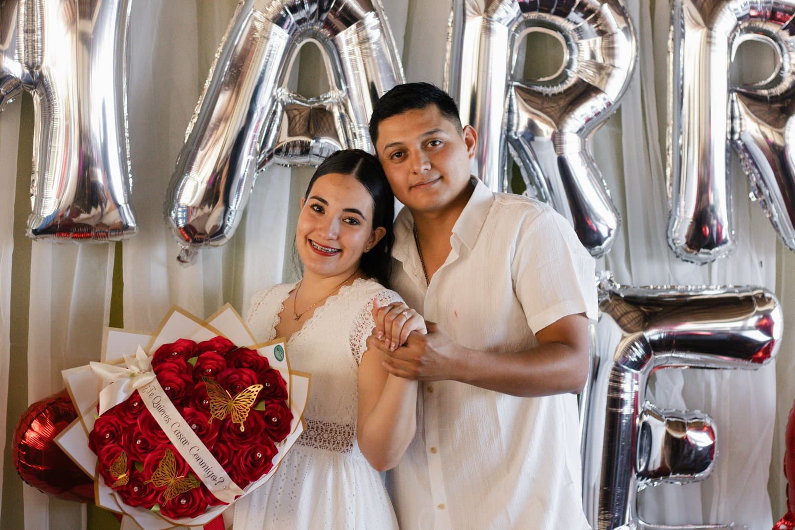 A newly engaged couple smile together in front of silver letter balloons, holding a box of red roses.