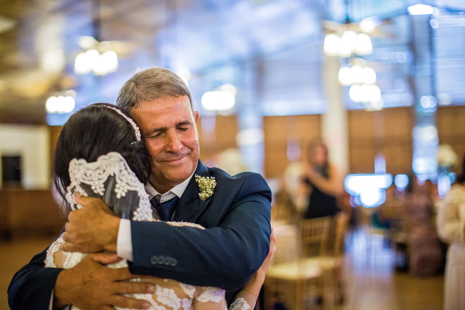 An older man in a navy suit and boutonniere closes his eyes as he holds the bride in a long hug.