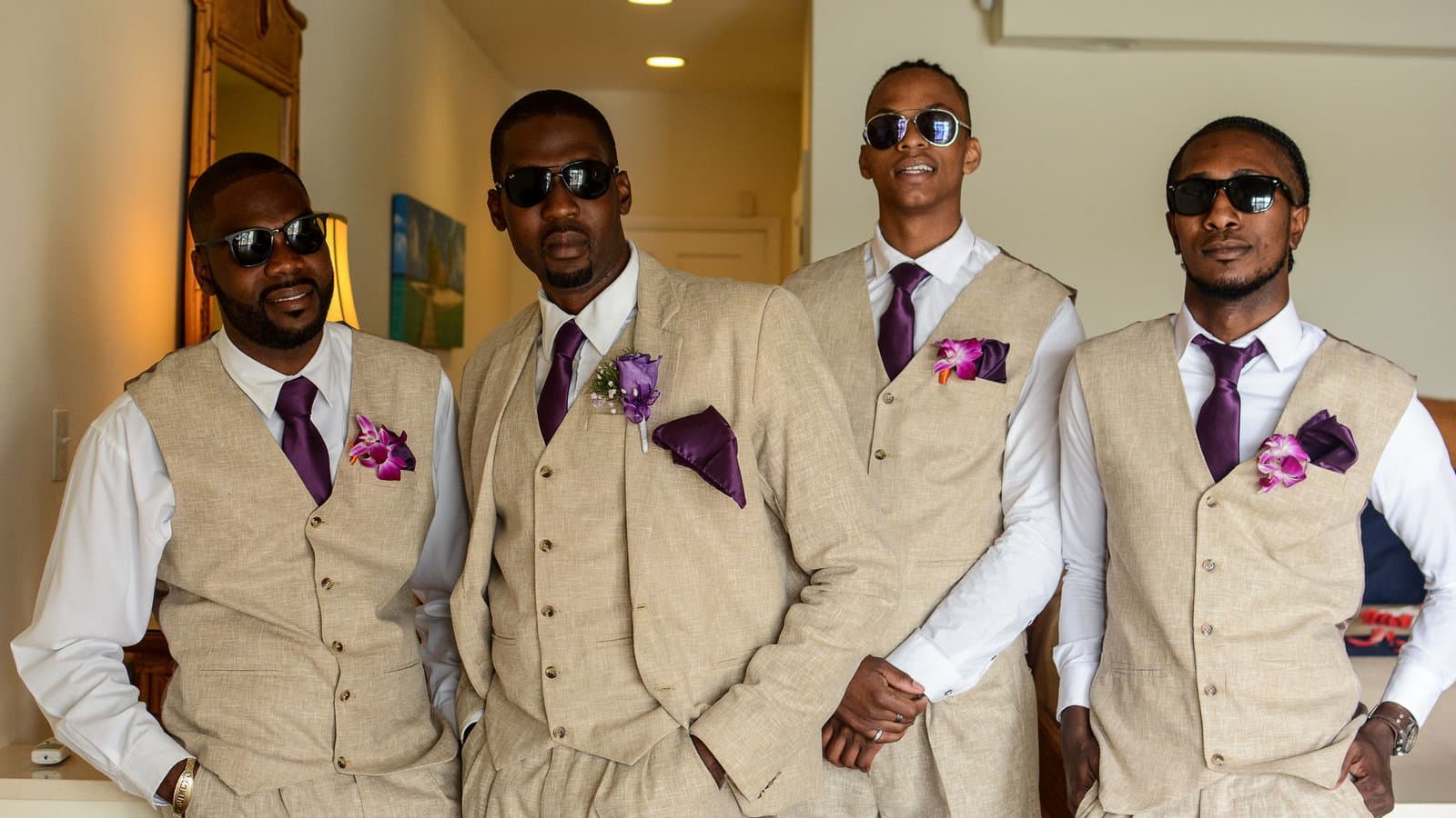 Four groomsmen in cream waistcoats with purple ties and boutonnieres stand together in daylight.