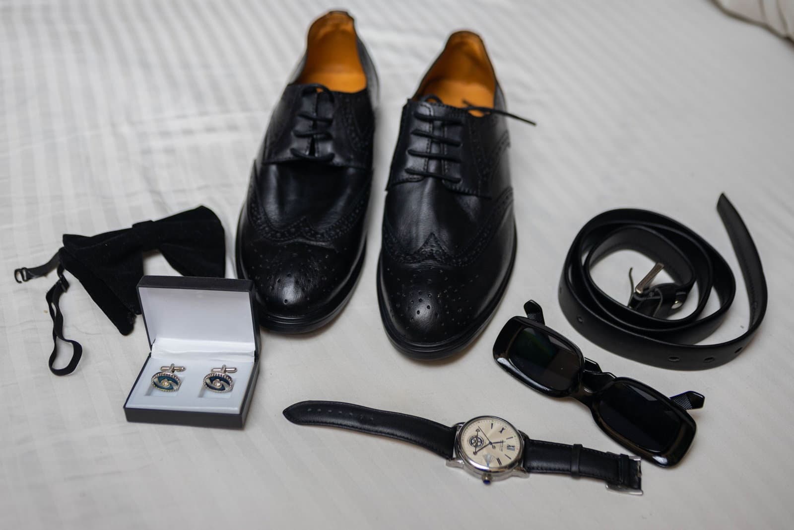 A flat lay of groomsmen gifts on white linen: leather brogues, cufflinks in a box, a watch, a belt and sunglasses.