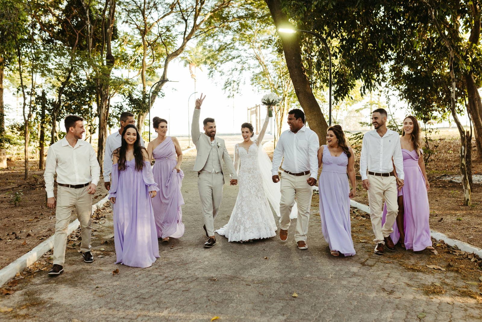 A wedding party of ten walks a tree-lined path in daylight, attendants in lilac gowns and cream suits either side of the couple.