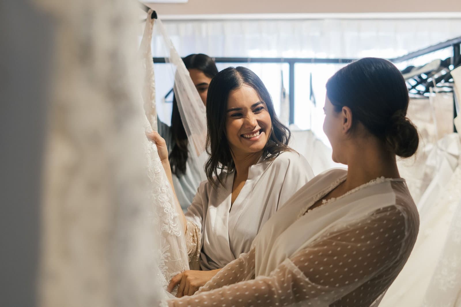 Two women smile together among hanging gowns in a bright bridal boutique during a dress appointment.
