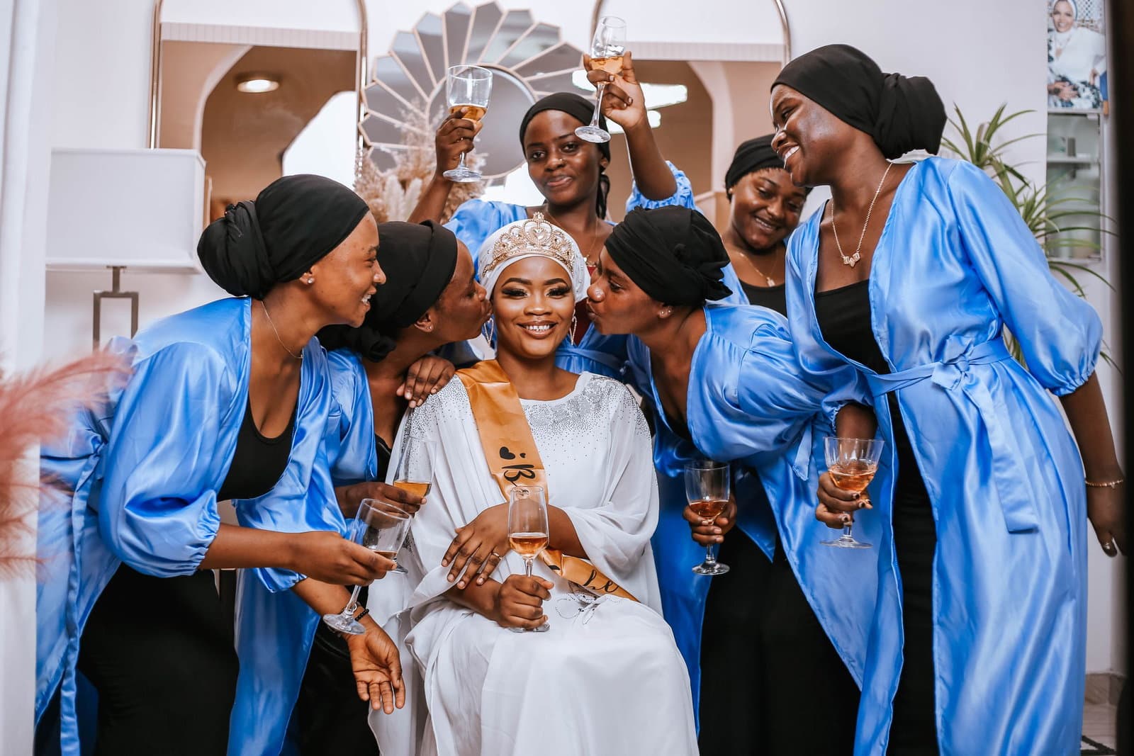 A bride surrounded by her wedding party in blue satin robes, raising glasses and laughing together.