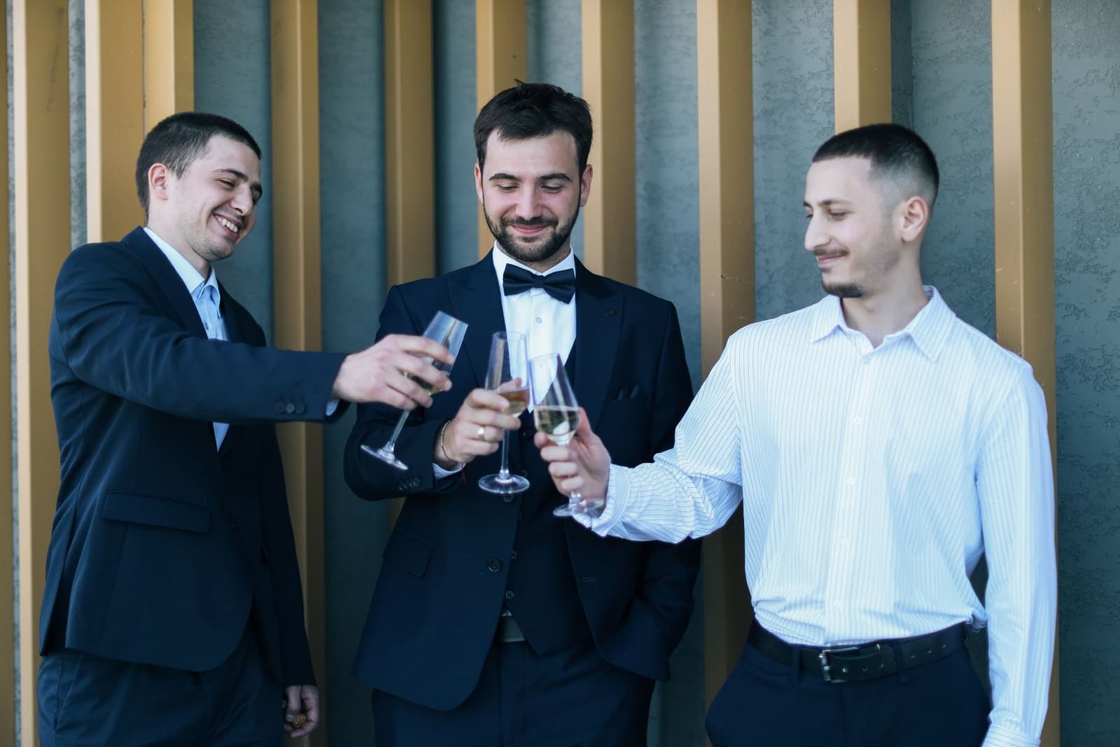 Three men in suits clink champagne glasses together outdoors in bright daylight.