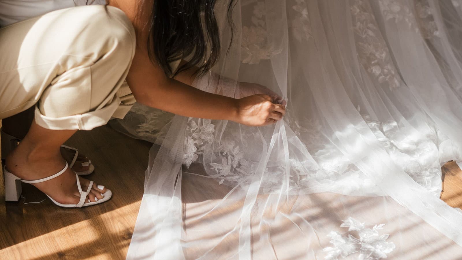 A maid of honor crouches down to straighten the hem of the bride's veil and gown in a sunlit room.