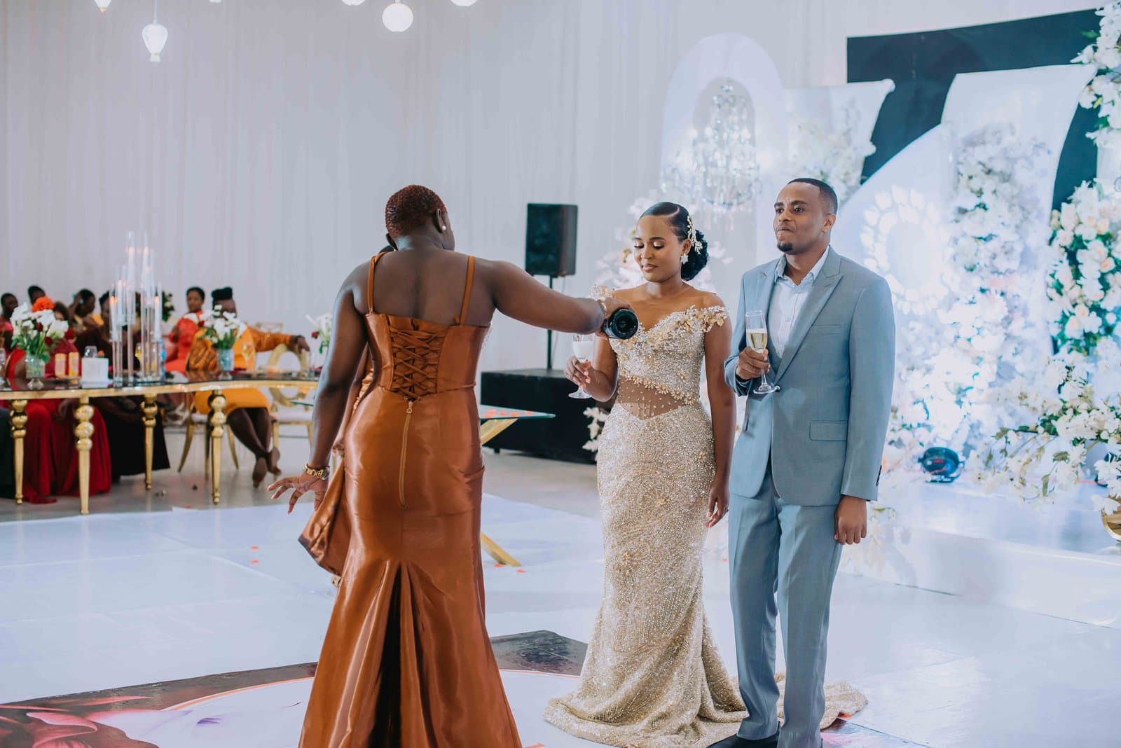An attendant in a rust satin gown raises a bottle toward the couple on a bright white reception floor as they hold their glasses.
