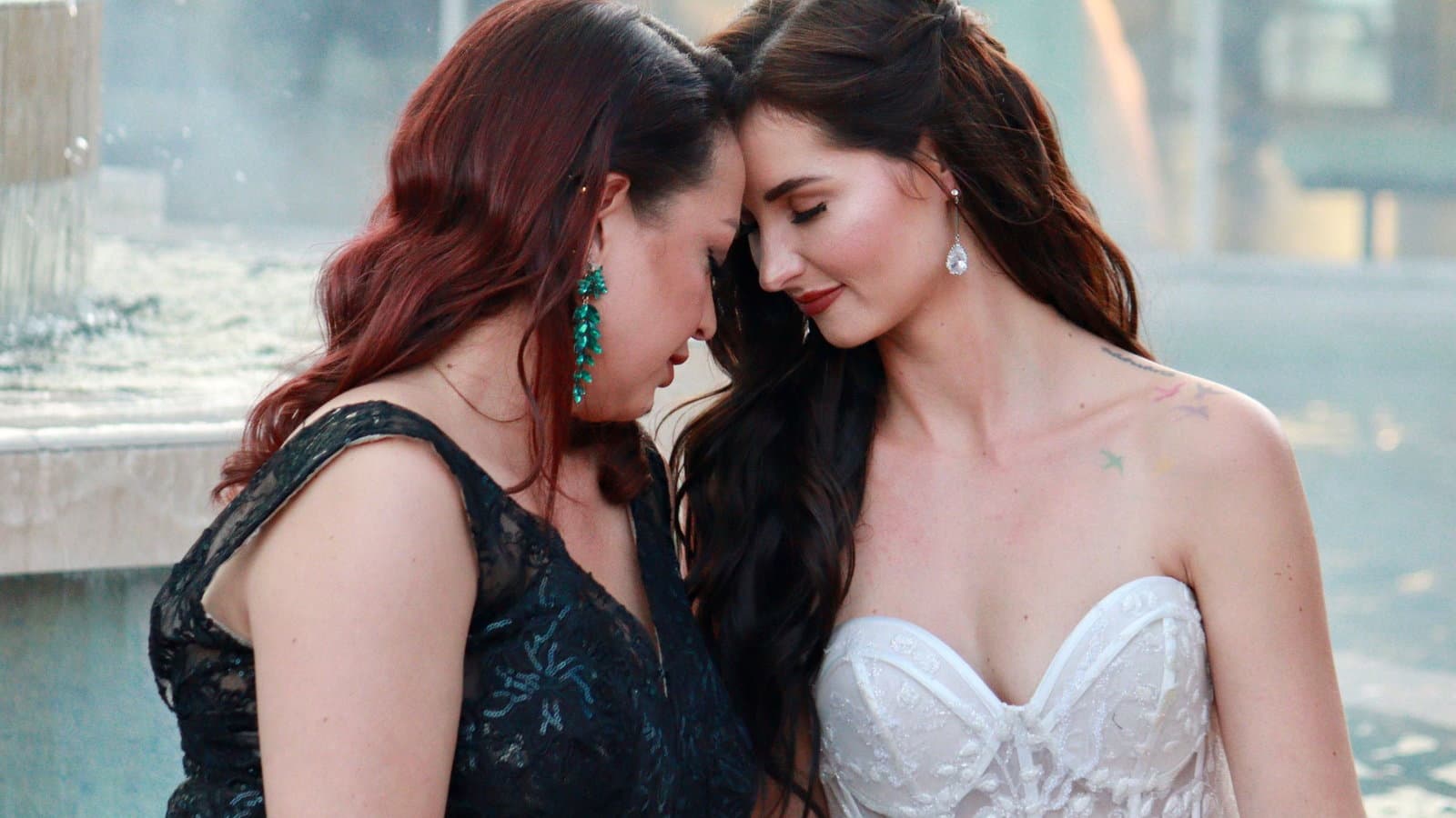 A bride in white and her attendant in a dark gown lean their foreheads together, smiling, in daylight.