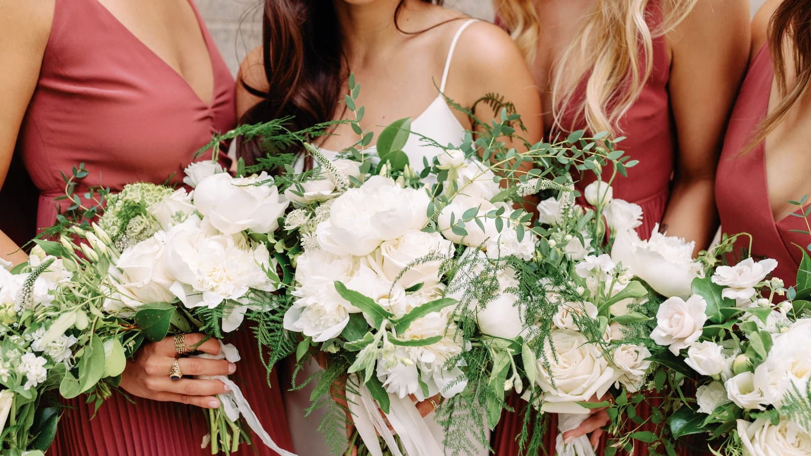 Bridesmaids in dusty rose dresses stand shoulder to shoulder holding white garden rose bouquets.