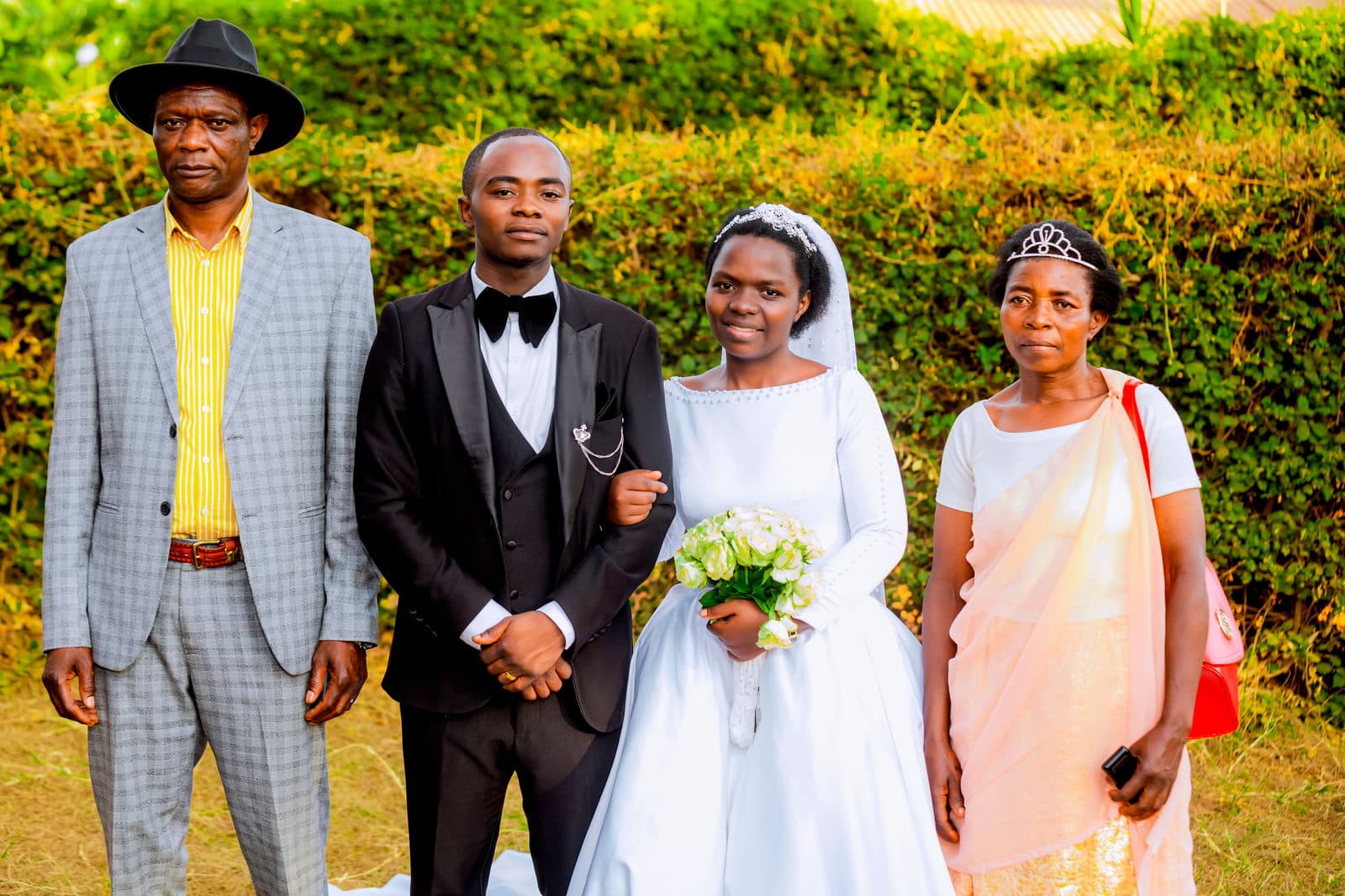 The groom's mother in a peach wrap stands beside the newlyweds and the groom's father in front of a sunlit hedge.