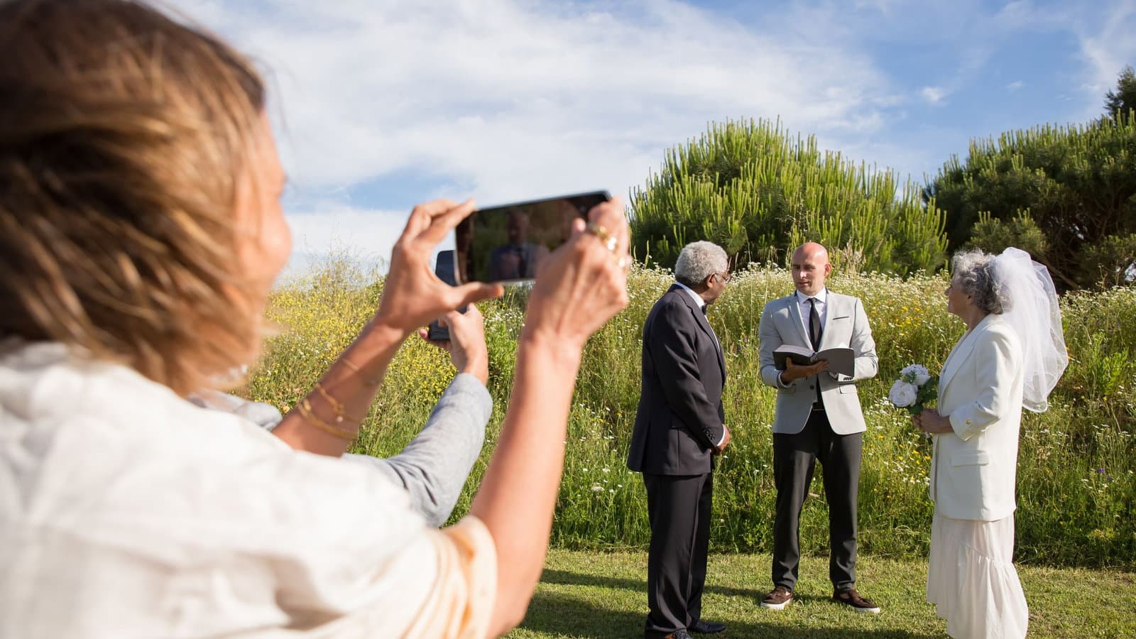A wedding guest films the couple and their officiant on her phone during an outdoor ceremony.