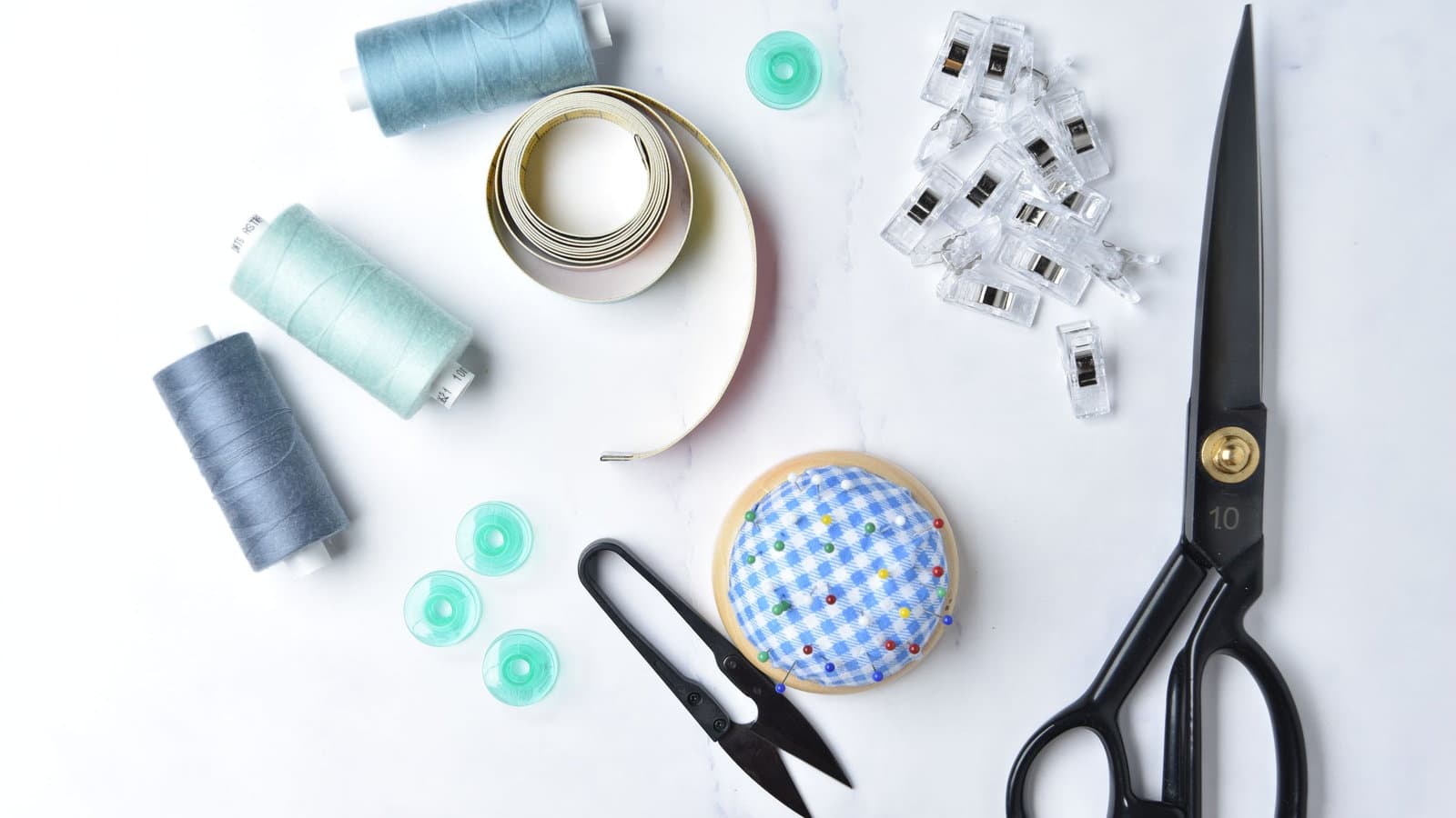 A flat lay of emergency kit essentials on white: thread spools, ribbon, fabric clips, pins and scissors.
