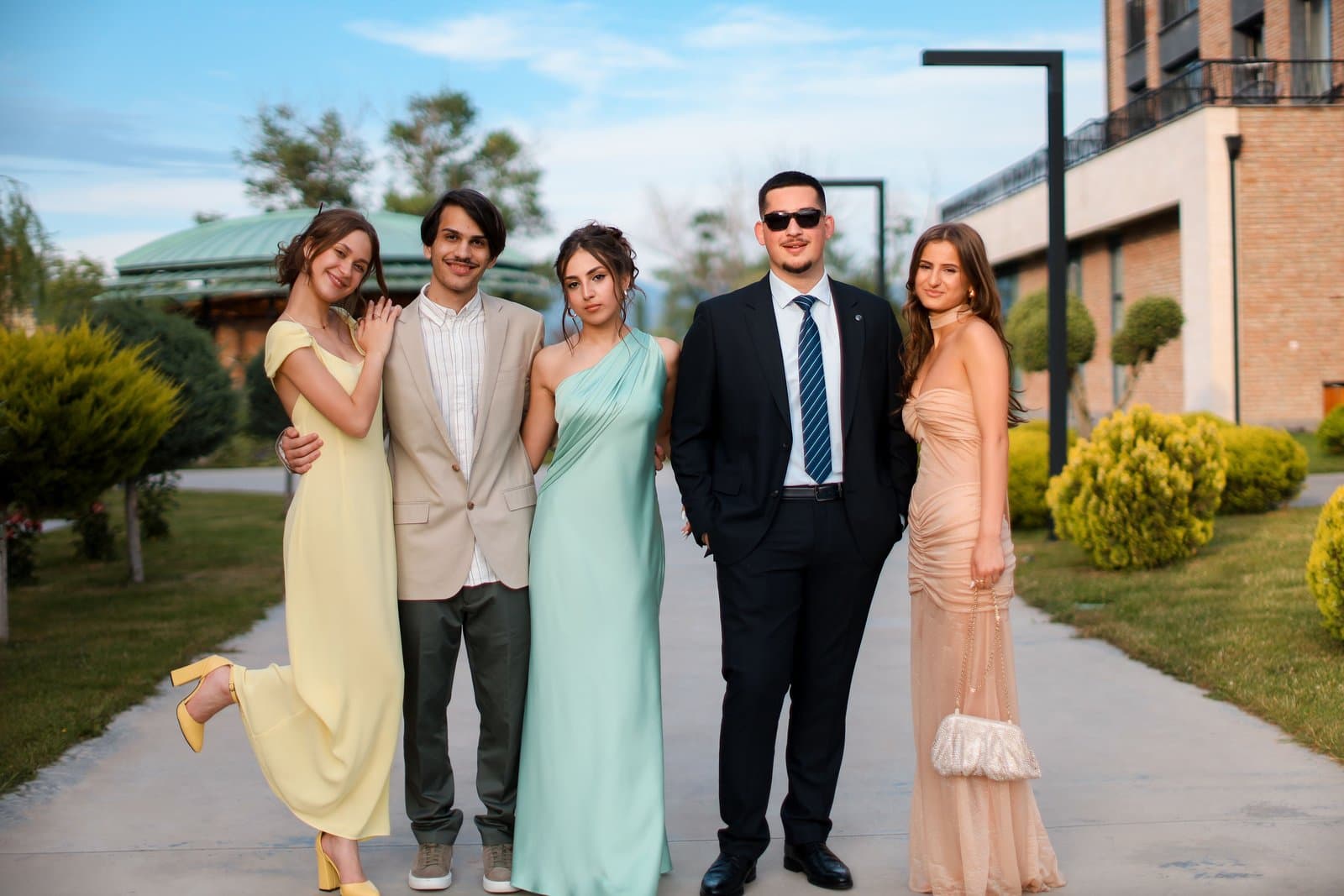 Five wedding guests in a mix of long gowns, a navy suit and a tan blazer walk a garden path in daylight.