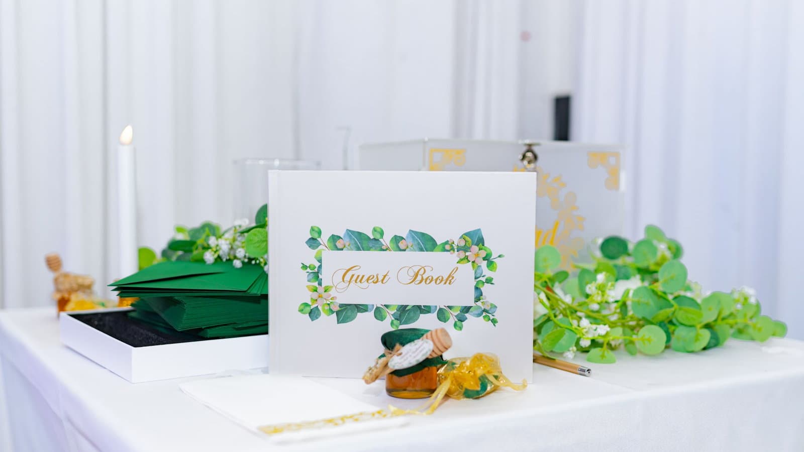 A floral-bordered wedding guest book open on a white draped table beside greenery and a candle.