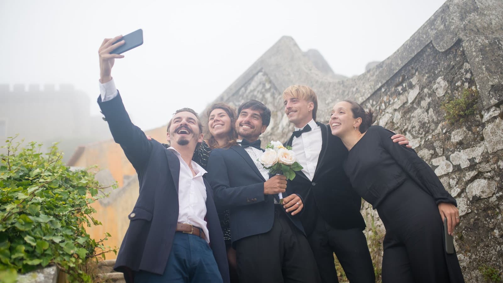 A wedding party of five laughs together as one of them holds a phone up for a group selfie outdoors.