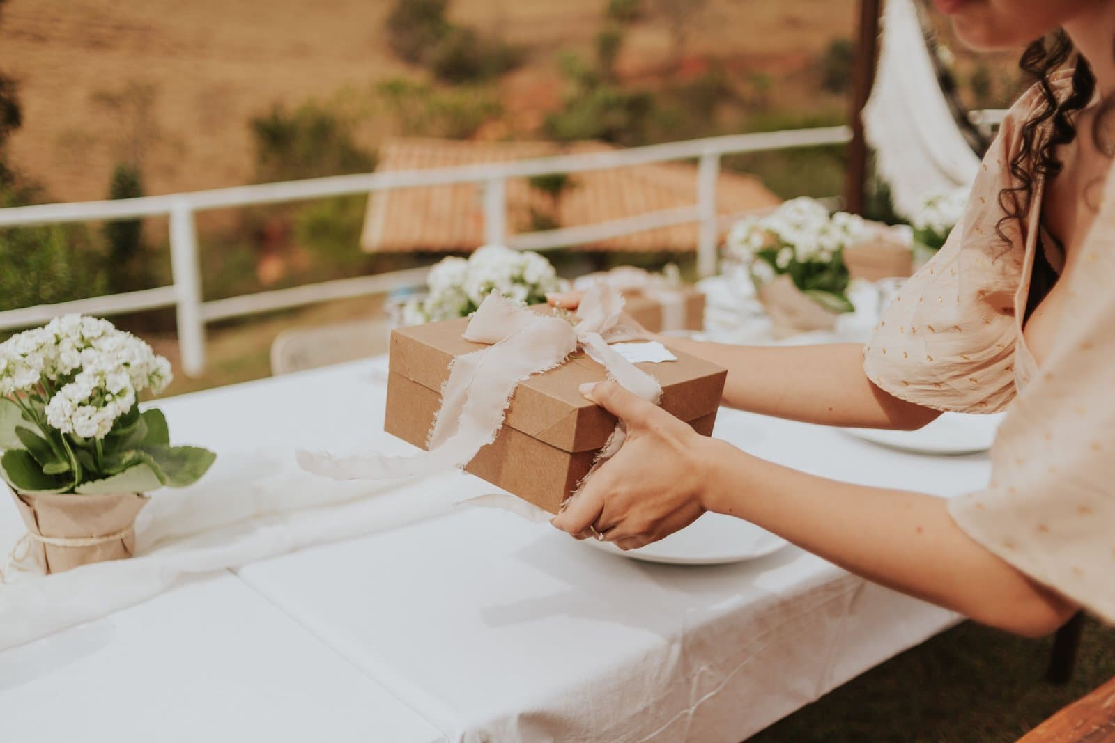 A woman sets a kraft gift box tied with a frayed silk ribbon onto a white-clothed table at an outdoor countryside wedding.