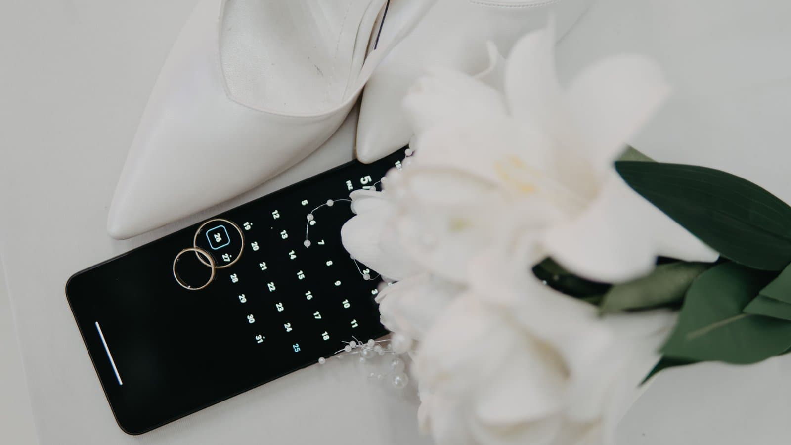 Two wedding rings rest on a phone showing a calendar with the date circled, beside white bridal shoes and fresh flowers.