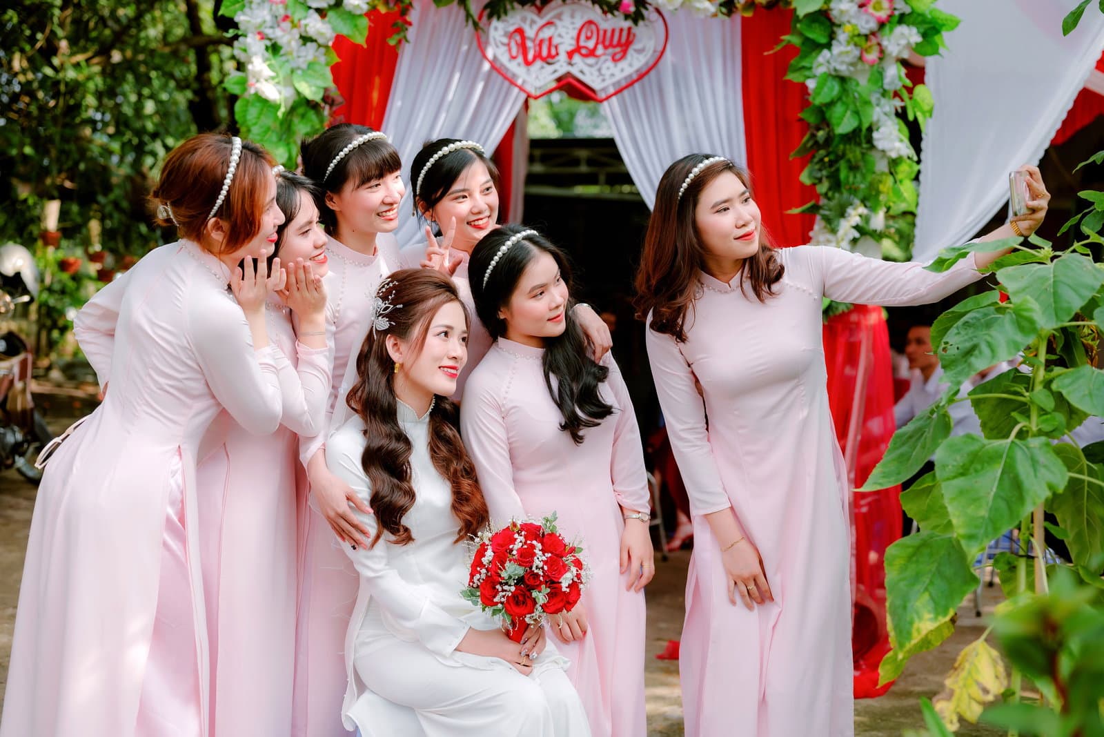 Bridesmaids in pink crowd around the seated bride for a group selfie held out at arm's length beside a flower-trimmed doorway.