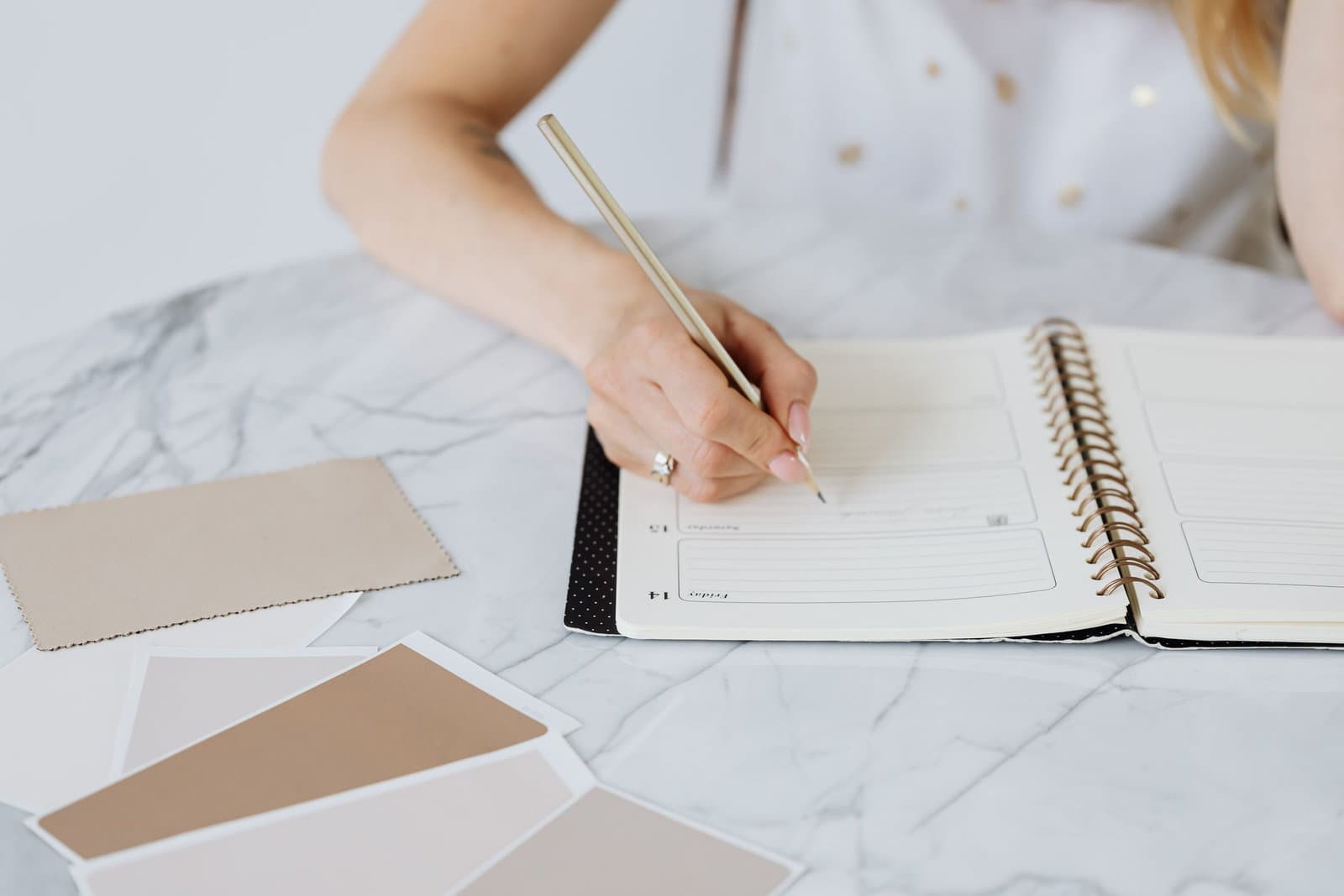 A hand writes into a dated planner on a marble desk beside neutral colour swatches.