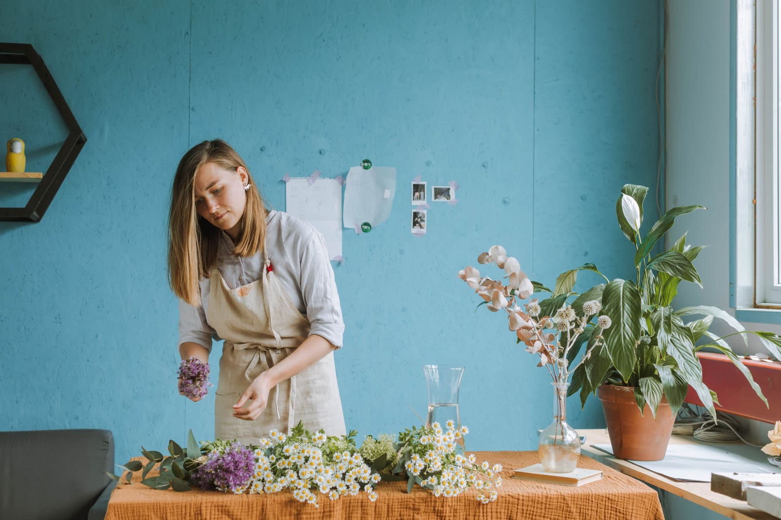 A florist in an apron arranges stems at a worktable in a bright studio full of plants.