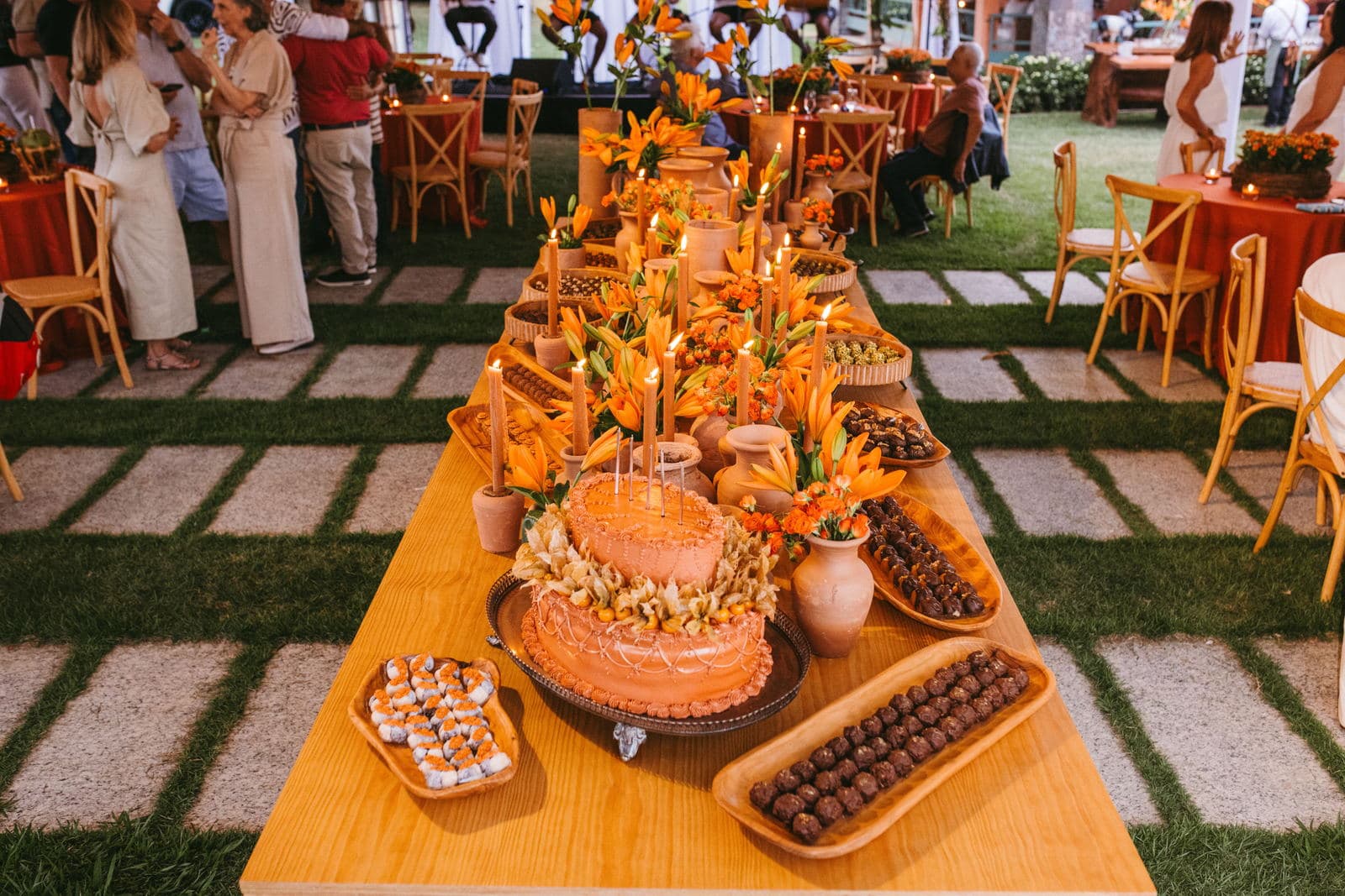 A long welcome table on a lawn laid with cake, sweets, candles and orange lilies, with guests gathered around it in daylight.