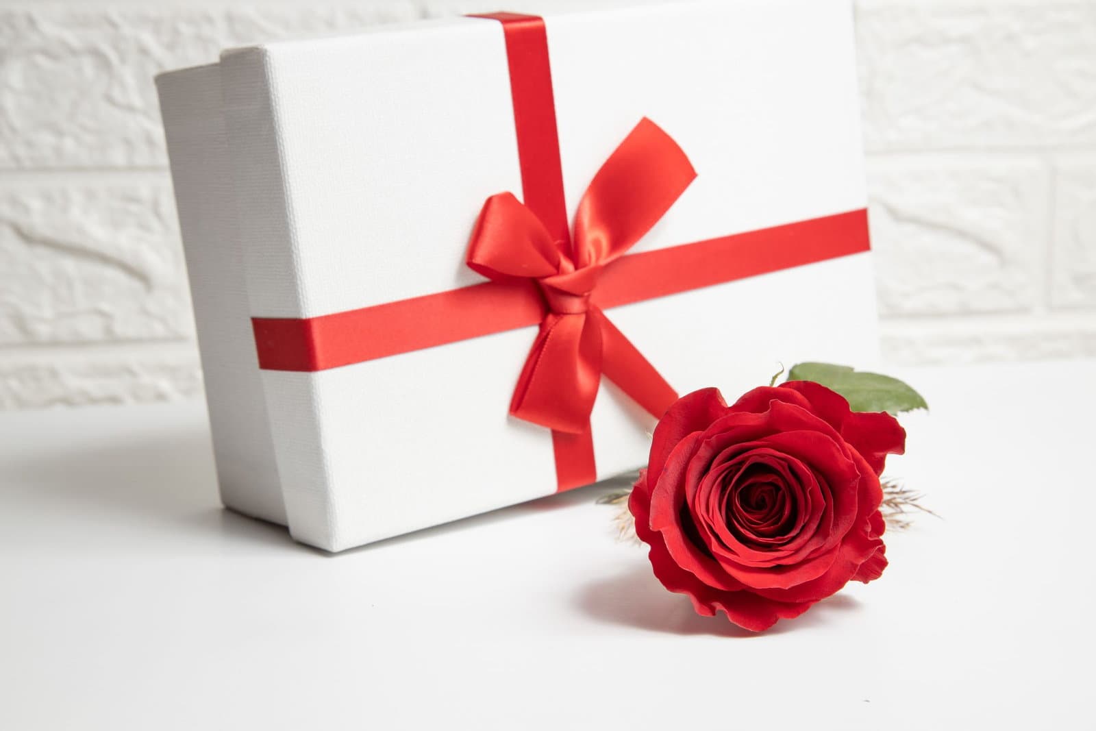 A white gift box tied with a red satin ribbon beside a single red rose on a bright surface.