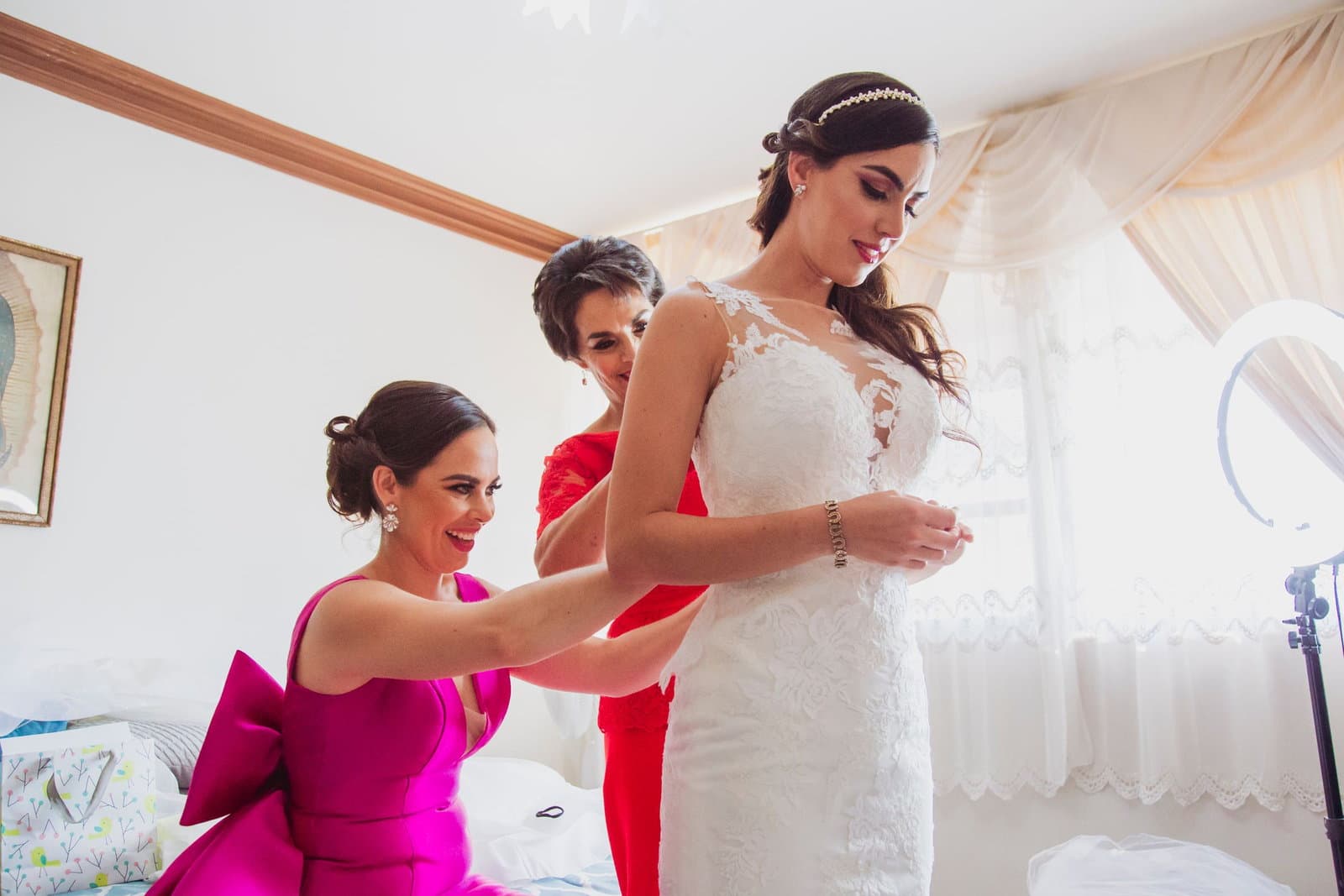 Two women in bright dresses help the bride fasten the back of her lace gown in a sunlit room.